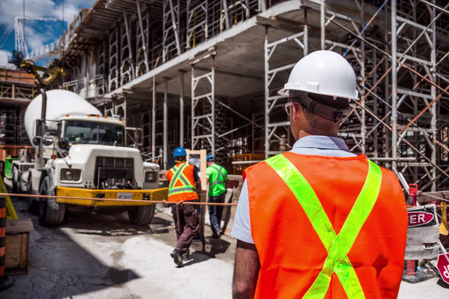 a construction labour supply worker stands in front of a large building under construction.He is wearing a hard hat and an orange safety vest. A truck is parked in front, and  several other workers are scattered around the area.